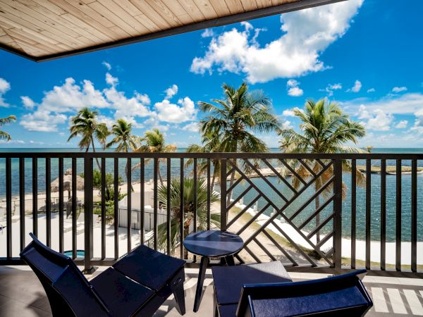 A balcony with two chairs and a small table, overlooking a tropical beach with palm trees and clear blue skies.
