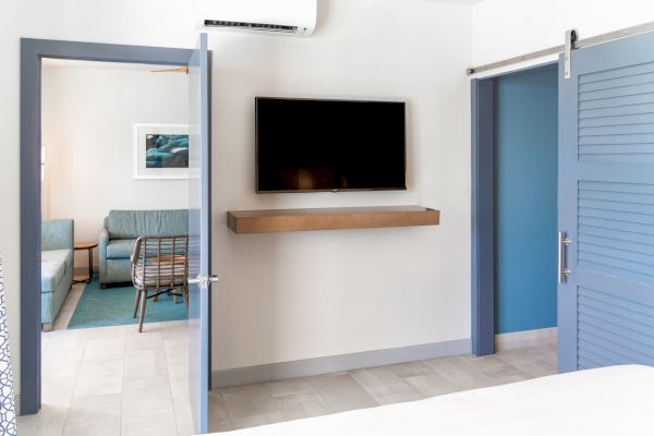 The image shows a modern, minimalist room with a wall-mounted TV, blue doors, and a visible adjacent living area with a sofa and wall art.