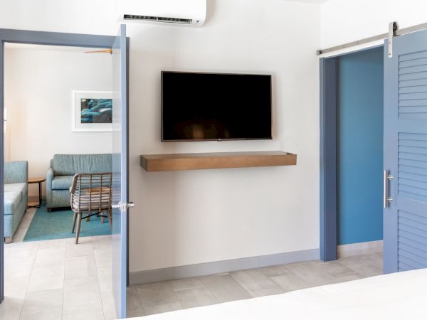 The image shows a modern, minimalist room with a wall-mounted TV, blue doors, and a visible adjacent living area with a sofa and wall art.