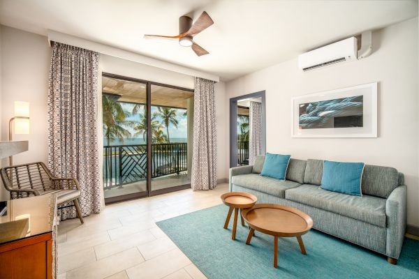 A modern, airy living room features a grey sofa with blue cushions, wooden tables, and a sliding glass door opening to a balcony with a sea view.