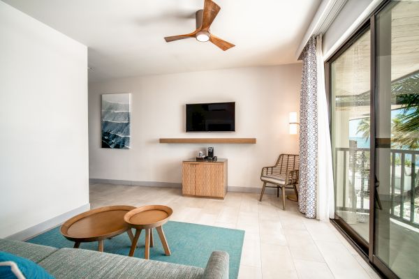 A modern living room with a TV, wall art, coffee tables, sofa, ceiling fan, large windows and glass doors opening to a balcony with a view.