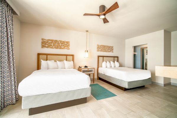 The image shows a hotel room with two beds, wall art, a bedside table, and a ceiling fan. The room has a bright, minimalistic design and tiled flooring.