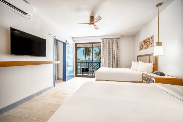 This image shows a hotel room with two beds, a wall-mounted TV, a balcony overlooking palm trees and the ocean, and modern decor.