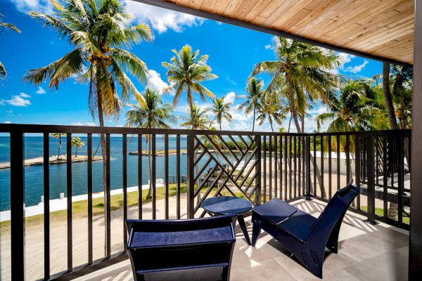 A tropical balcony with chairs and a table overlooks palm trees and a serene blue ocean under a bright, sunny sky.