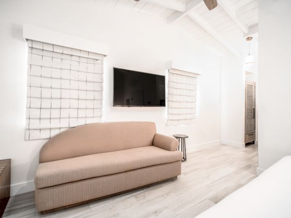 A modern living area with a beige couch, wall-mounted TV, and two windows with white blinds; features wooden flooring and a minimalist design.