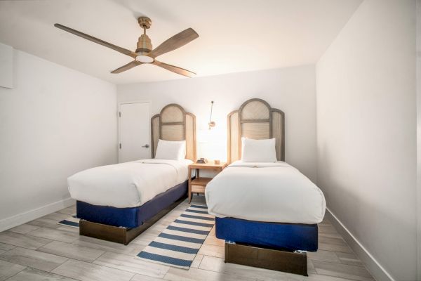 The image shows a room with two single beds, a ceiling fan, striped rug, and a shared nightstand, all set against a minimalist white backdrop.