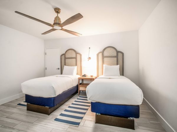 The image shows a room with two single beds, a ceiling fan, striped rug, and a shared nightstand, all set against a minimalist white backdrop.