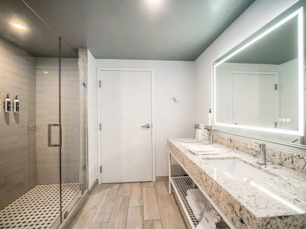 Modern bathroom with a glass-enclosed shower, large vanity, dual sinks, and a backlit mirror on a light wood floor.
