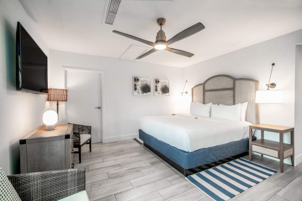 The image shows a modern hotel room with a large bed, ceiling fan, TV, and stylish decor in neutral tones.