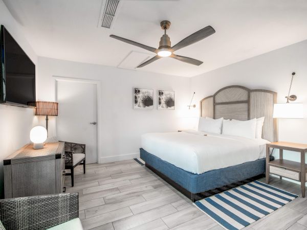 The image shows a modern hotel room with a large bed, ceiling fan, TV, and stylish decor in neutral tones.