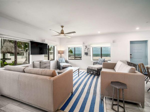 A modern living room with sofas, a blue-striped rug, a ceiling fan, and large windows offering a beach view.