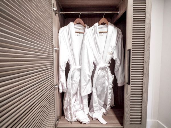 Two white bathrobes hanging inside a wooden closet on hangers, neatly arranged with the closet door partially open.