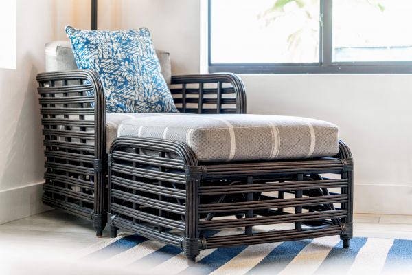 A black wicker armchair with a gray cushion and blue patterned pillow beside a window on a striped rug.