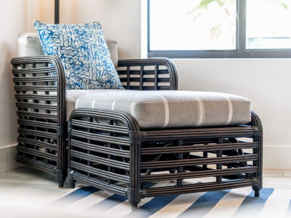 A black wicker armchair with a gray cushion and blue patterned pillow beside a window on a striped rug.
