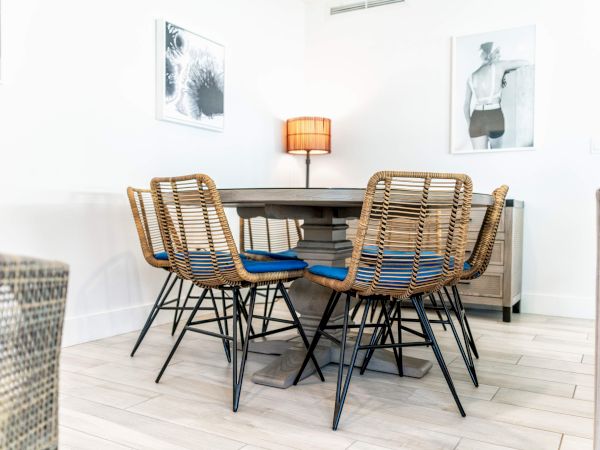 A dining area with a round table, six wicker chairs, a central lamp, and two wall art pieces on a light wooden floor.