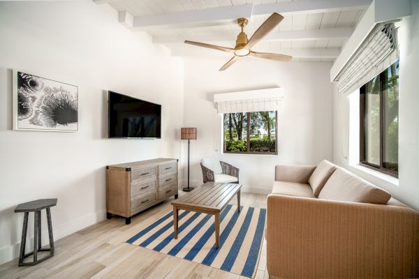 A modern living room with white walls, a beige couch, striped rug, coffee table, flat-screen TV, ceiling fan, artwork, and large windows.