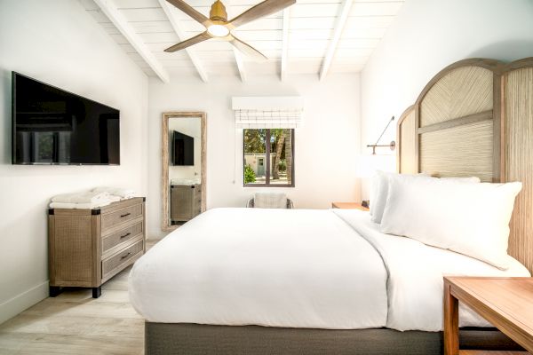 The image shows a modern, clean bedroom with a large bed, a ceiling fan, a wall-mounted TV, and a window with a view of trees outside.