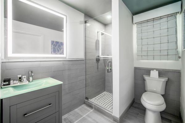 A modern bathroom featuring a lighted mirror, vanity, glass-enclosed shower, and toilet with a window above it.