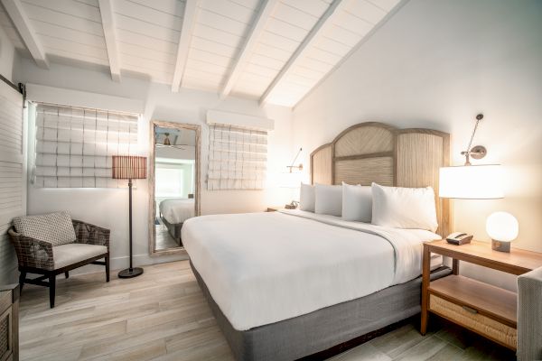 A cozy bedroom featuring a large bed, wicker chair, bedside table with lamp, standing mirror, and wall-mounted decor, all in a soft color scheme.