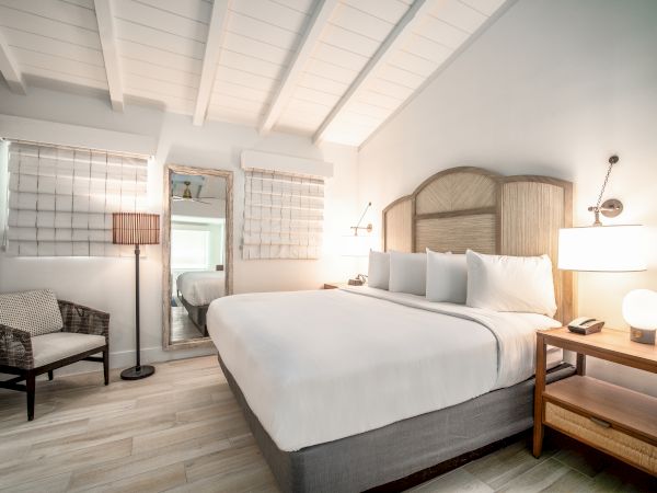 A cozy bedroom featuring a large bed, wicker chair, bedside table with lamp, standing mirror, and wall-mounted decor, all in a soft color scheme.