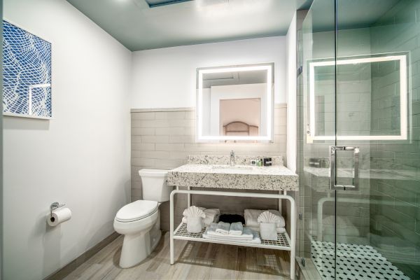 A modern bathroom with a toilet, granite countertop sink, backlit mirror, glass shower, and wall art, featuring a towel rack below the sink.