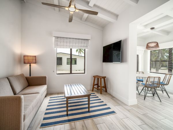 A modern living room with a sofa, striped rug, and a wall-mounted TV, leading to a dining area with a table and chairs under pendant lights.
