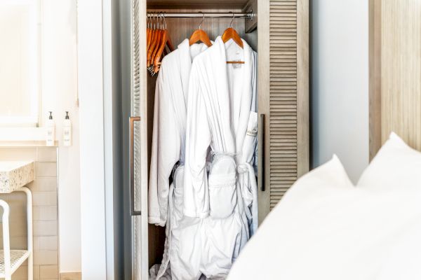 The image shows a closet containing several white bathrobes hanging on wooden hangers, next to a bed with white linens in a bright room.