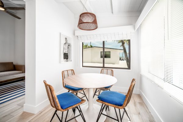 A small dining area with a round table and four blue-cushioned chairs, featuring a window, wall art, and a woven hanging lamp, is shown.