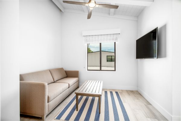 A small room with a beige sofa, striped rug, wooden coffee table, wall-mounted TV, window, and ceiling fan, all in white walls and light wood flooring.