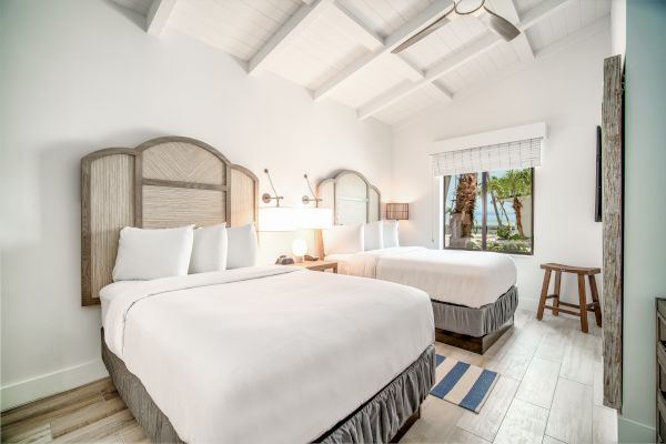 A bright, cozy bedroom with two double beds, white bedding, wooden headboards, a window with a view, and light wooden flooring.