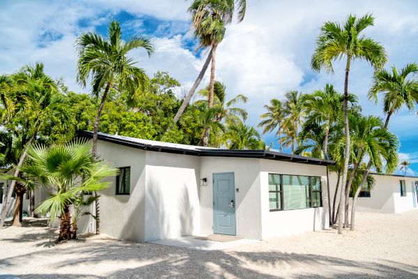 A small, white building with a light blue door, surrounded by palm trees and clear skies, giving a tropical and serene feel to the setting.