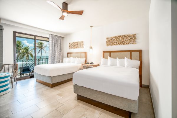 The image shows a bright, modern hotel room with two large beds, a ceiling fan, balcony with ocean view, and tropical decor.