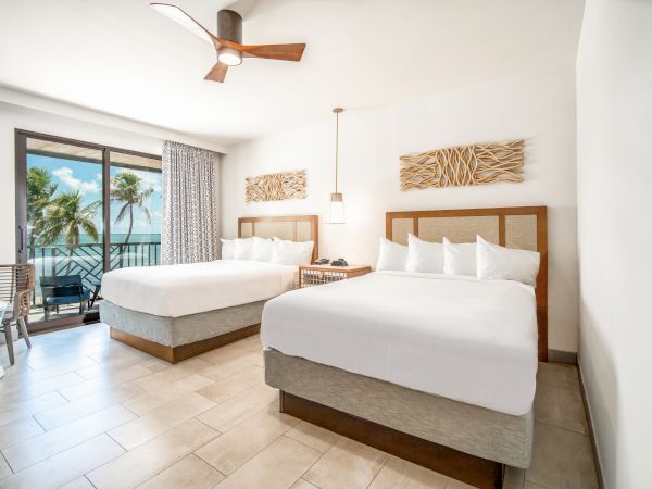 The image shows a bright, modern hotel room with two large beds, a ceiling fan, balcony with ocean view, and tropical decor.