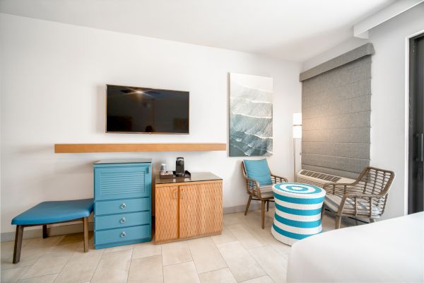 A modern hotel room with a wall-mounted TV, a blue bench, a cabinet with a coffee maker, a painting, chairs with a round striped table, and a window.