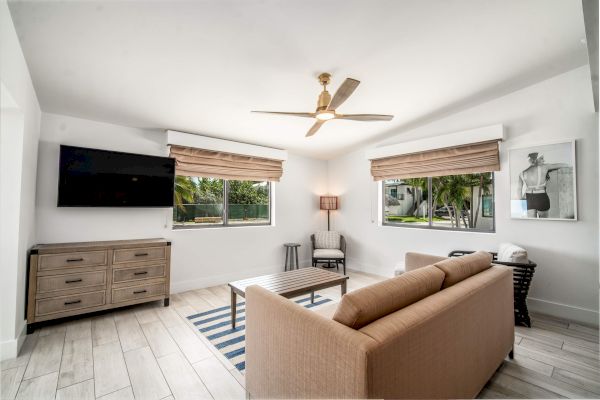 A modern living room with a couch, striped rug, wall-mounted TV, wooden furniture, and two windows with blinds, featuring a ceiling fan.