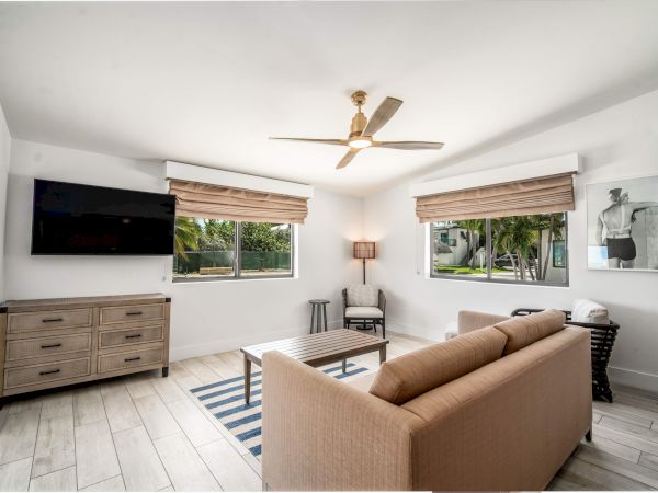A modern living room with a couch, striped rug, wall-mounted TV, wooden furniture, and two windows with blinds, featuring a ceiling fan.