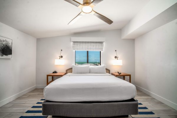 The image shows a modern bedroom with a large bed, two side tables, lamps, a ceiling fan, and a window with a view, in a minimalistic style.