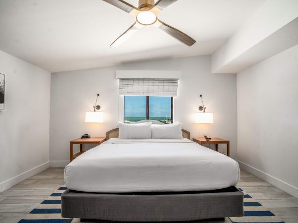 The image shows a modern bedroom with a large bed, two side tables, lamps, a ceiling fan, and a window with a view, in a minimalistic style.