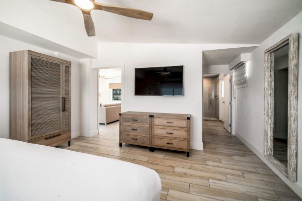 A bedroom with wooden furniture, a mounted TV, ceiling fan, and mirrored closet. Light color scheme with tiled flooring.