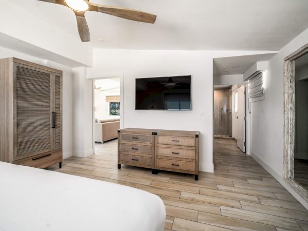 A bedroom with wooden furniture, a mounted TV, ceiling fan, and mirrored closet. Light color scheme with tiled flooring.