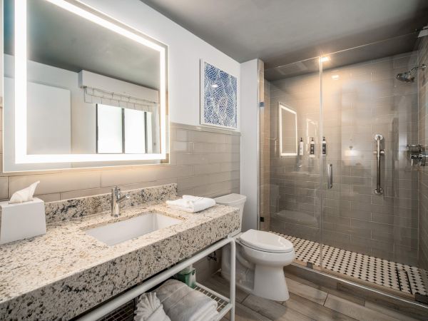 A modern bathroom with a granite-topped vanity, illuminated mirror, glass shower, and neatly organized towels and toiletries.