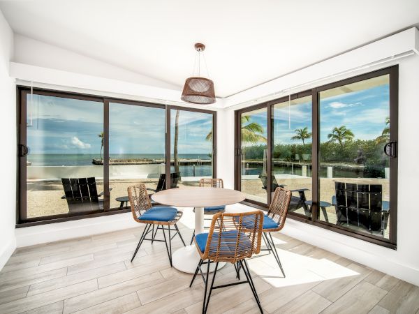 A bright, modern dining area with large windows offering a view of a tropical outdoor scene, featuring a round table and four chairs.