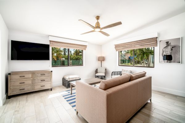 This image shows a modern, bright living room with a flat-screen TV, light brown sofa, wooden dresser, coffee table, wall art, and large windows.