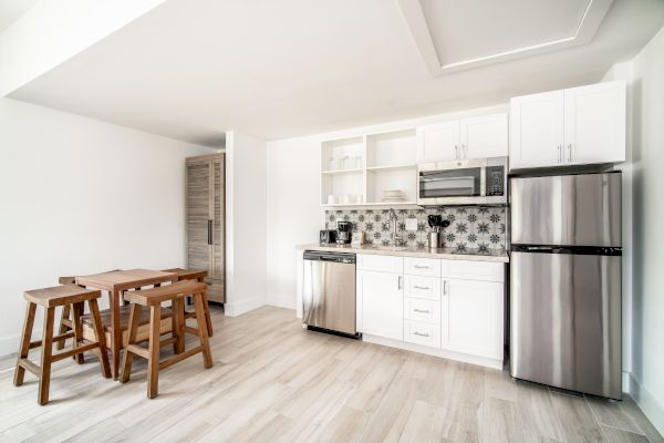 A modern kitchen with white cabinets, stainless steel appliances, a wooden dining table with stools, and light wood flooring.