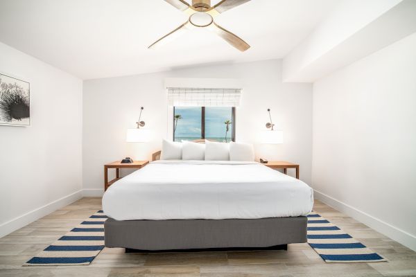 A minimalist bedroom with a large bed, bedside tables, a ceiling fan, wall art, window, and striped rugs beneath the bed on a wooden floor.