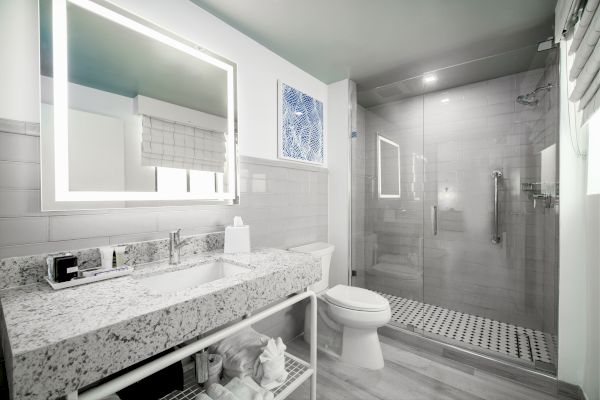 A modern bathroom with a granite countertop, large mirror, toilet, and glass-enclosed shower. Shelving below the sink holds towels and toiletries.