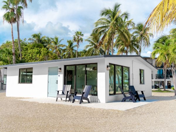 A small white house with large windows, surrounded by palm trees, featuring outdoor seating on a gravel yard under a clear blue sky.