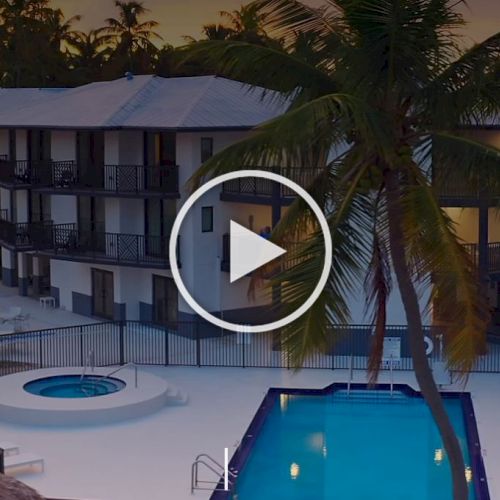 A pool and hot tub in front of a multi-story building with balconies, surrounded by palm trees at dusk.