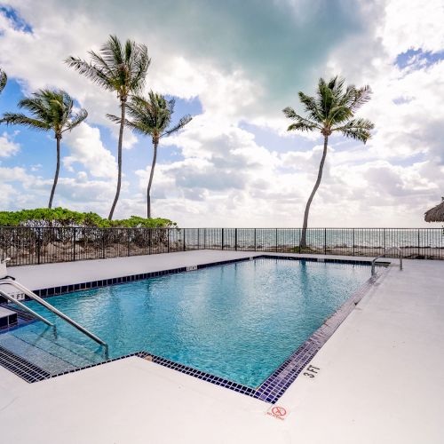 A pool surrounded by palm trees with ocean views, sun umbrellas, and blue sky with clouds in the background.