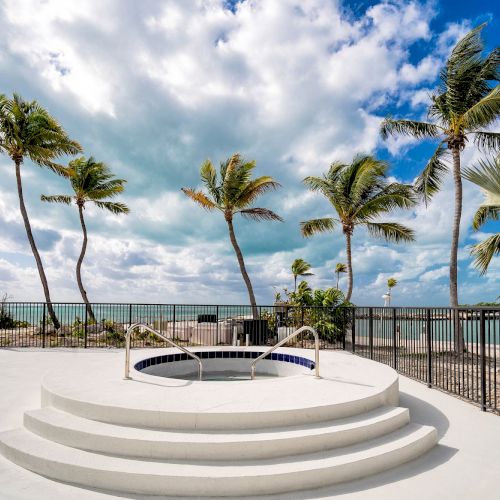 A tropical beachfront scene with palm trees, a circular hot tub on a white platform, and ocean views under a partly cloudy sky.